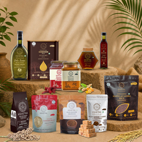 A collection of Kshetra brand organic food products arranged on rustic wooden blocks against a warm beige backdrop with decorative leaves and grains. The products include olive oil, sesame seeds, honey, chutney, jam, spices, and candies.